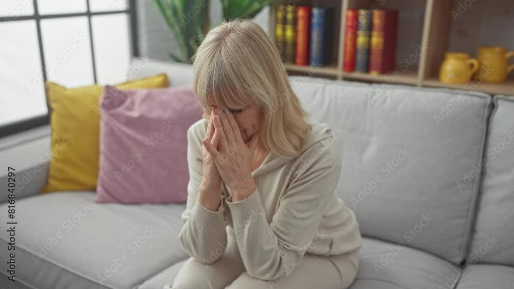A distressed blonde woman sits on sofa in her home, conveying emotions of worry or sadness. A distressed blonde woman sits on sofa in her home, conveying emotions of worry or sadness.