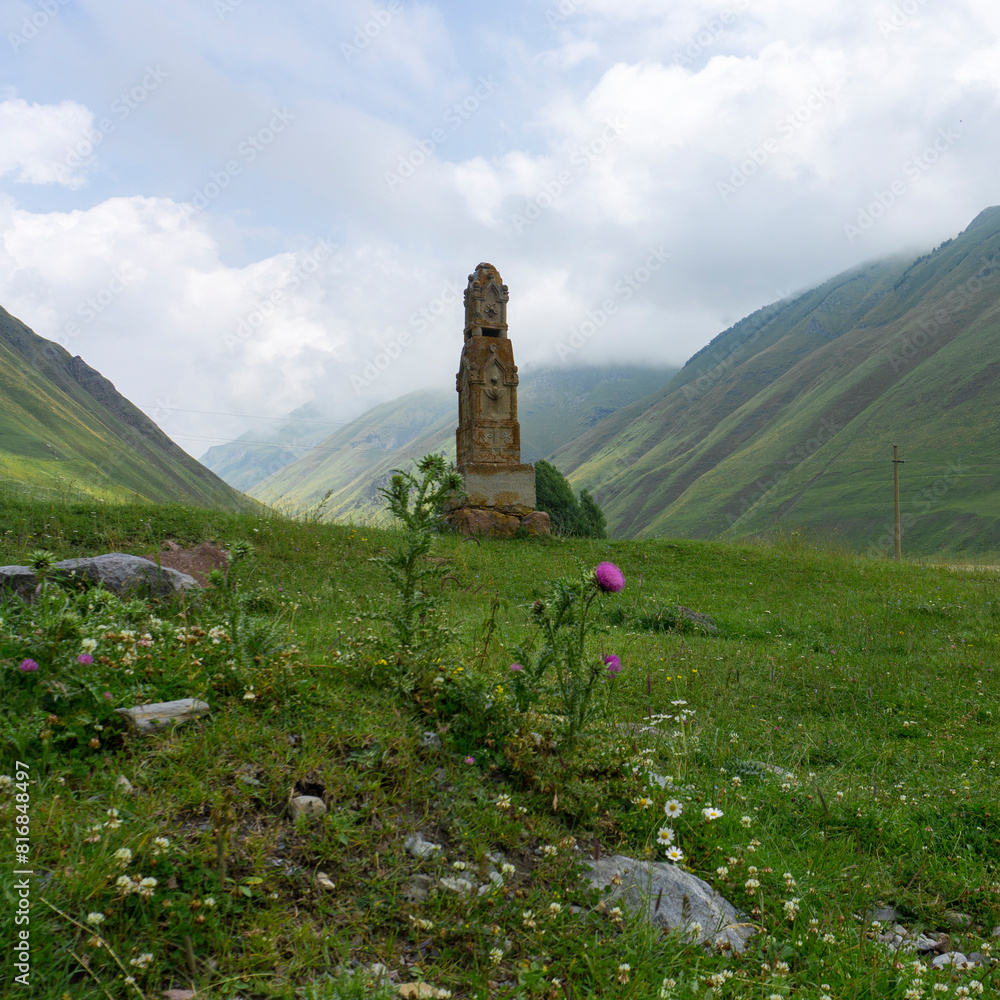 Stone pedestal with a cross on top in a gorge among the mountains. Grass, stones and purple flower