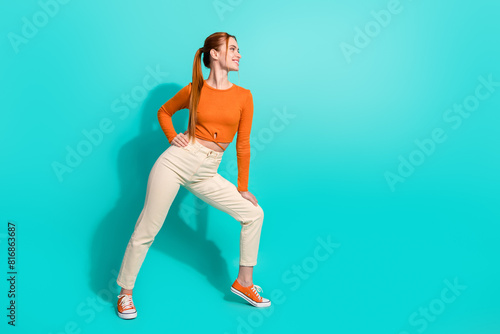 Full size photo of positive girl with ginger hair dressed orange shirt look at promo empty space isolated on teal color background