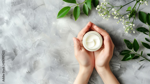 Hands holding a jar of white cream on a grey textured background, surrounded by green leaves and white flowers, suggesting a natural and organic skincare product. © Natalia