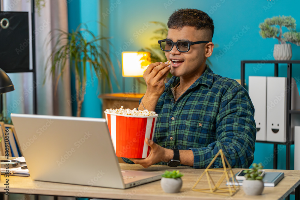 Business Indian man freelancer taking break from work wearing 3D glasses eating popcorn watching movie on laptop. Smiling Arabian guy sitting at home office desk having snacks during break. Lifestyle Business Indian man freelancer taking break from work wearing 3D glasses eating popcorn watching movie on laptop. Smiling Arabian guy sitting at home office desk having snacks during break. Lifestyle