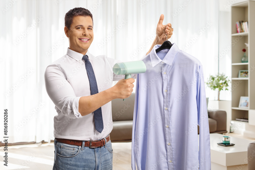 Obraz premium Happy young man using a hand steam iron for a shirt at home