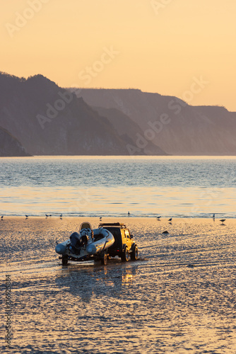 At low tide, an off-road car carries an inflatable rubber boat along the shallows to the sea. Morning seascape at sunrise. Beautiful view of the sea bay and mountains. Golden morning sunlight at dawn.
