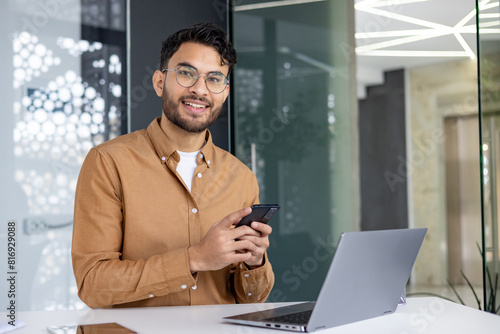 Smiling young professional using smartphone and laptop in modern office environment
