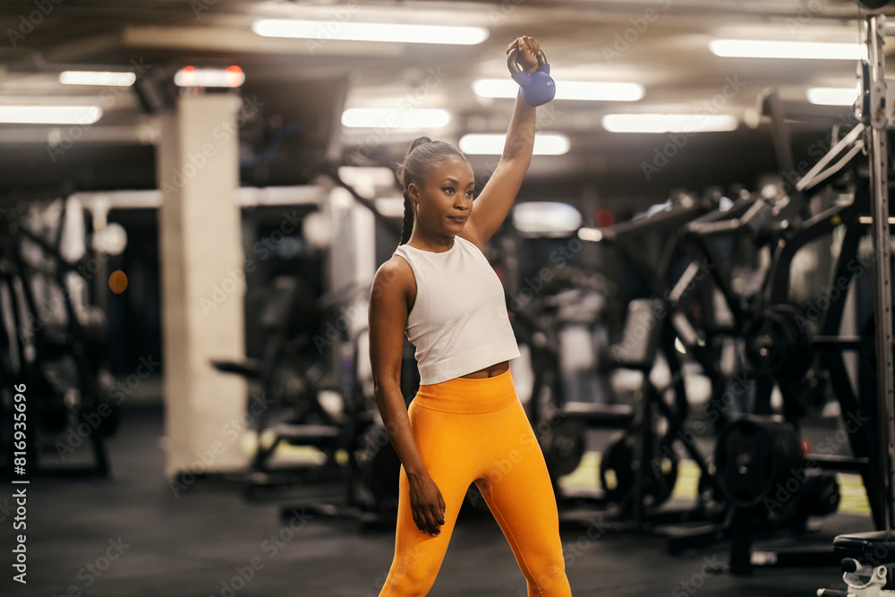 A black sportswoman in shape is practicing her shoulder with kettle bell at the gym.