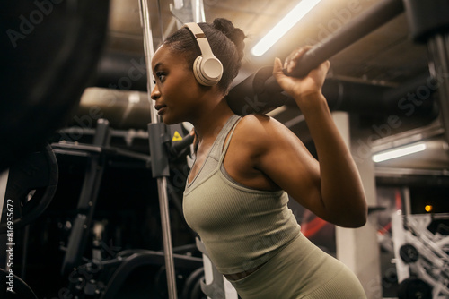 Behang Strong black sportswoman with headphones squatting doing workouts on smith machine at gym