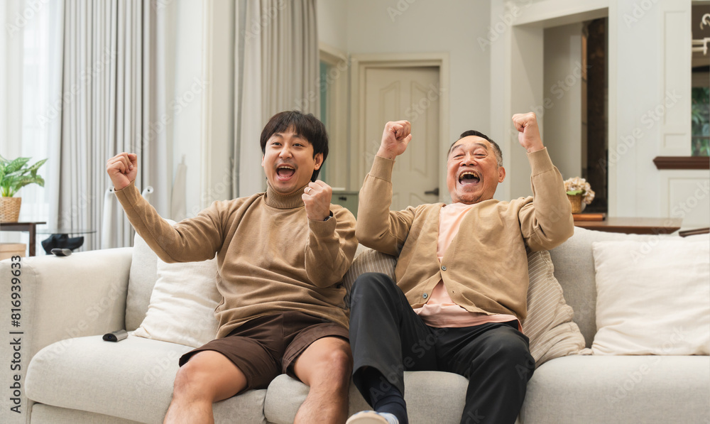 Obraz premium Cheerful Asian old man and middle aged son enjoy watching tv, celebrating good news. Smiling two male generations family feeling excited together, raising hands, resting on sofa in living room at home
