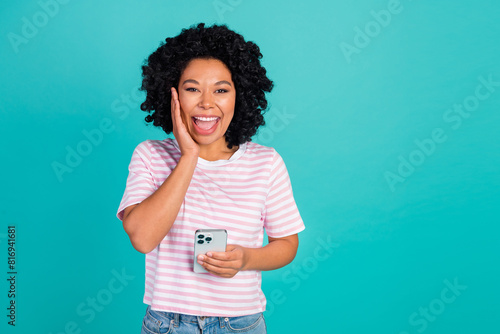 Portrait photo of young happy girl touch cheek wearing striped t shirt using phone surprised isolated on aquamarine color background