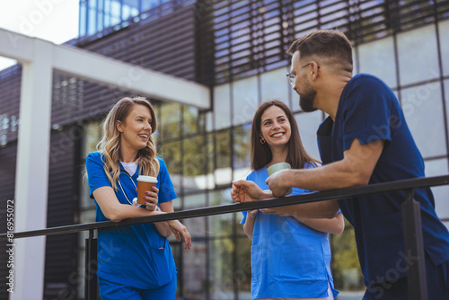 Fotografie Two female and one male healthcare worker in blue scrubs share a light-hearted moment with coffee cups in hand, standing outside a medical facility