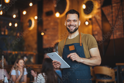 Принти на полотні A cheerful male waiter, clad in an apron, holds a digital tablet while serving customers in a bustling bar environment