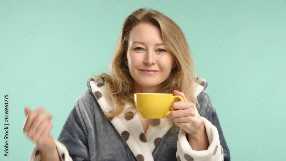Captured on a calm turquoise background, a young woman savors the inviting aroma of fresh coffee, holding a yellow cup, dressed in a cozy polka dot robe. Captured on a calm turquoise background, a young woman savors the inviting aroma of fresh coffee, holding a yellow cup, dressed in a cozy polka dot robe.
