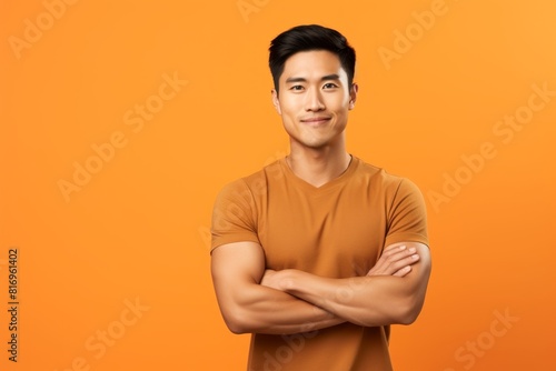 Portrait of a content asian man in his 20s with arms crossed while standing against soft orange background