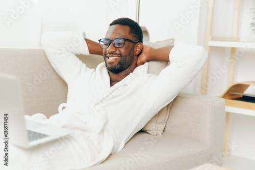 Smiling African American Freelancer Working on Laptop in Bathrobe, Sitting on Modern Sofa in Home Office Young, happy man typing on his computer in the morning, surrounded by cyberspace and technology