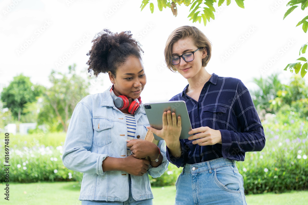 Fototapeta premium Two women are looking at a tablet together. One of them is wearing a blue shirt with a plaid pattern