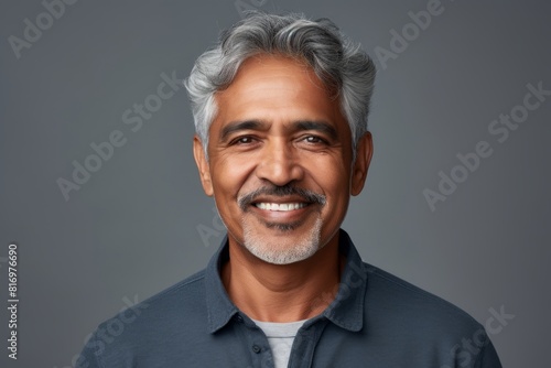 Portrait of a satisfied indian man in his 60s smiling at the camera while standing against soft gray background