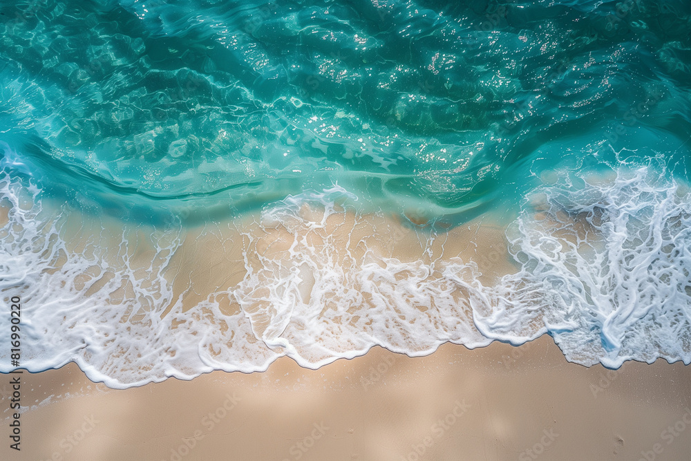 Fototapeta premium A top view of the crystal-clear azure sea, white wave foam, and beautiful sandy beach on a sunny day