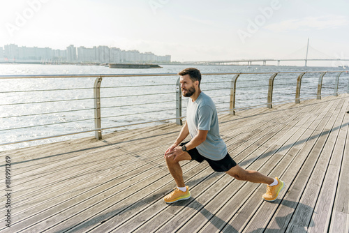 A man performs a lunge stretch on a wooden boardwalk by the water, with a bridge and city skyline in the background. He is dressed in athletic attire, preparing for exercise in the sunny weather.