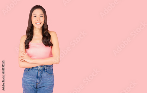 Portrait of a smiling young woman standing against a pink background, wearing casual clothes with arms crossed, exuding confidence and cheerfulness.


