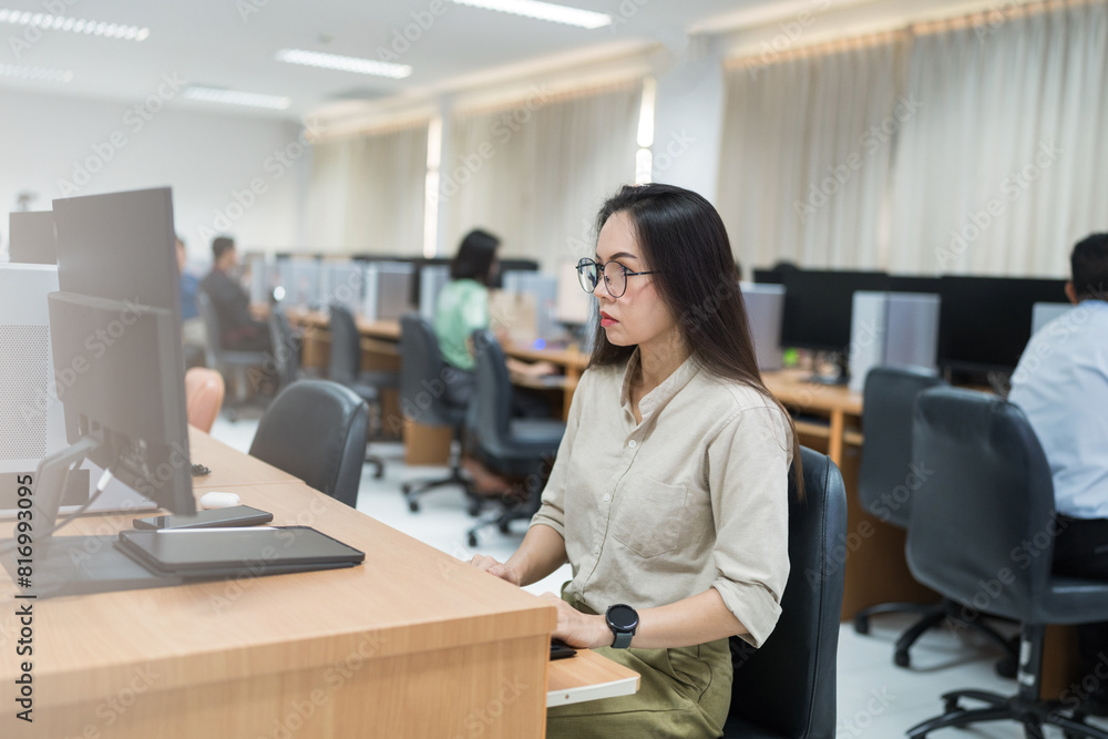 A woman sits at a desk in front of a computer. She is wearing glasses and is focused on her work. The room is filled with other people working on their computers, creating a busy A woman sits at a desk in front of a computer. She is wearing glasses and is focused on her work. The room is filled with other people working on their computers, creating a busy