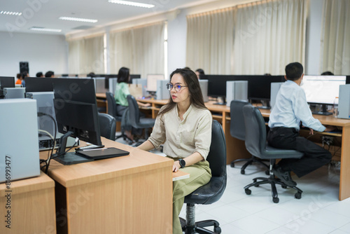 A woman sits at a desk in front of a computer. She is wearing glasses and is focused on her work. The room is filled with other people working on their computers, creating a busy