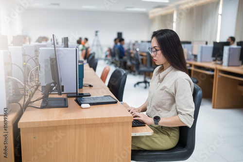 A woman sits at a desk in front of a computer. She is wearing glasses and is focused on her work. The room is filled with other people working on their computers, creating a busy