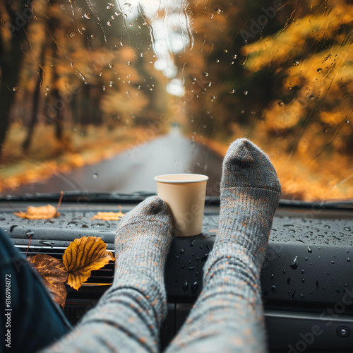 
A woman's legs in cozy socks rest on a car dashboard as she enjoys warm tea during a autumn journey, rain droplets decorating the windshield