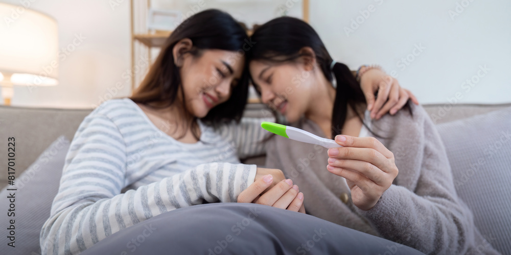 Fototapeta premium Couple smiling while looking at a positive pregnancy test on the couch at home. Women sharing a joyful moment anticipating parenthood together