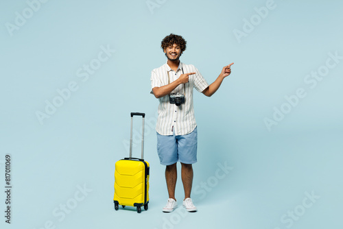 Full body traveler Indian man wear white casual clothes hold bag point aside isolated on plain blue background. Tourist travel abroad in free spare time rest getaway. Air flight trip journey concept. photo