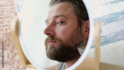 Reflection of a young bearded man shaving his beard holding electric shaver in bathroom. Handsome man sitting in front of the mirror. 30s male cutting trimming mustache using trimmer. Morning routine photo