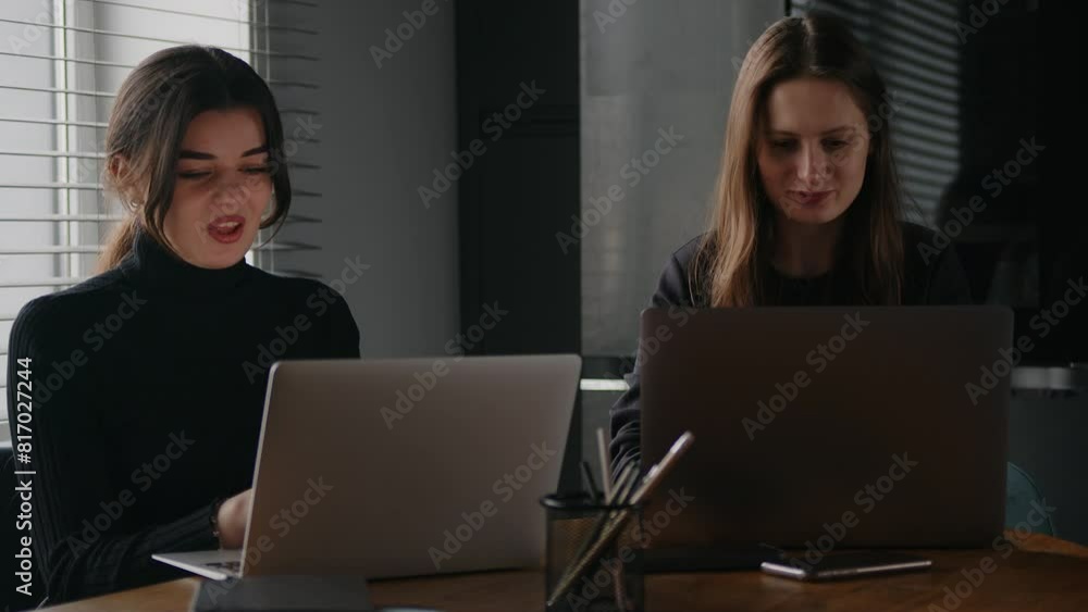 Two young women sitting in the office, working on laptops and talking to each other. Two young women sitting in the office, working on laptops and talking to each other.