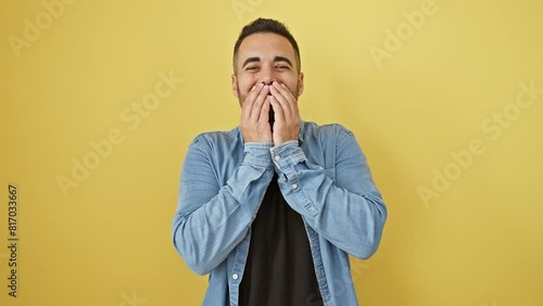 Young hispanic guy in denim shirt caught in gossip, laughing and covering mouth, posed against yellow isolated background, expressing embarrassed joy and shame photo
