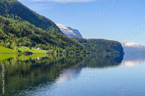 A tranquil Norwegian lake mirrors the lush green forests and distant snow-capped peaks under a clear blue sky  creating a stunning and peaceful scene.