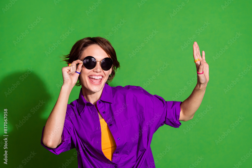 Photo portrait of young positive lady with bob hair wearing stylish sunglass dancing hipster raised arm up isolated on green color background Photo portrait of young positive lady with bob hair wearing stylish sunglass dancing hipster raised arm up isolated on green color background