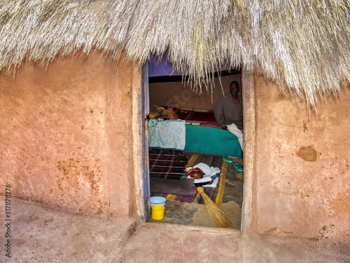 african mud house with thatched roof; man with baby inside of a shack; african poverty in the village