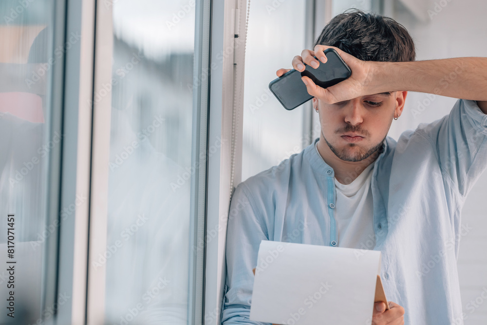 Fototapeta premium young man with mobile phone indoors next to the window with expression of stress or surprise