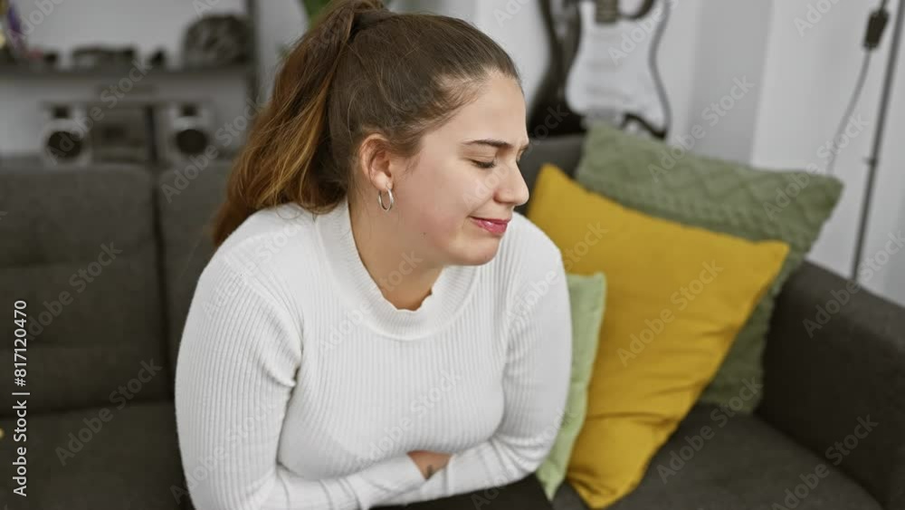 A young hispanic woman grimaces in pain, holding her abdomen while sitting on a grey couch indoors. A young hispanic woman grimaces in pain, holding her abdomen while sitting on a grey couch indoors.