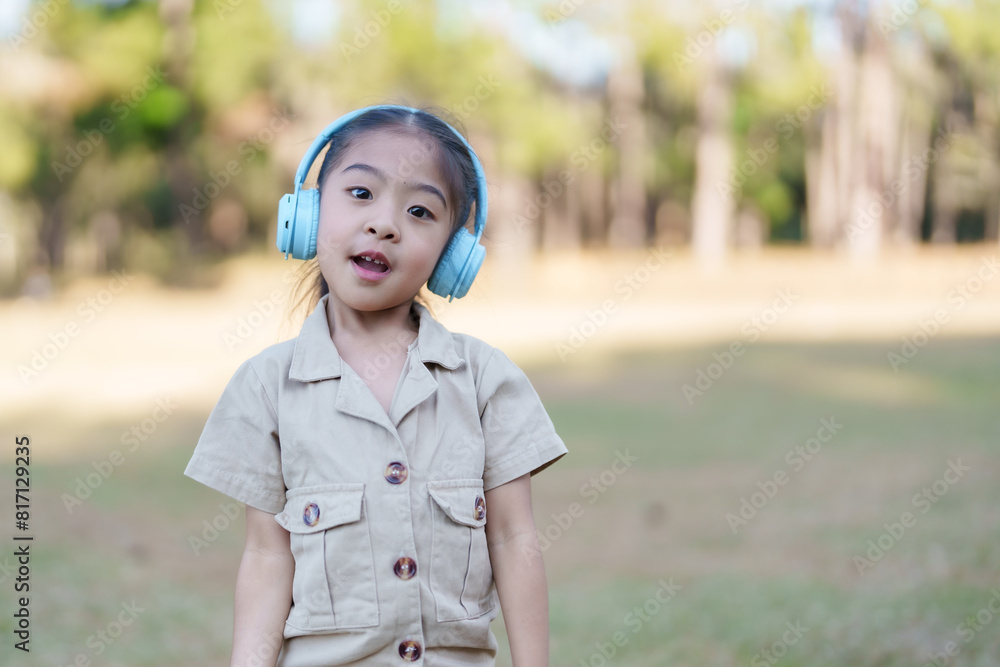 Asian girl blue headphones beige outfit standing park. Bright sunny day green trees. Expressive face enjoying music outdoors. Happy playful moment in nature. Carefree childhood scene. Asian girl blue headphones beige outfit standing park. Bright sunny day green trees. Expressive face enjoying music outdoors. Happy playful moment in nature. Carefree childhood scene.