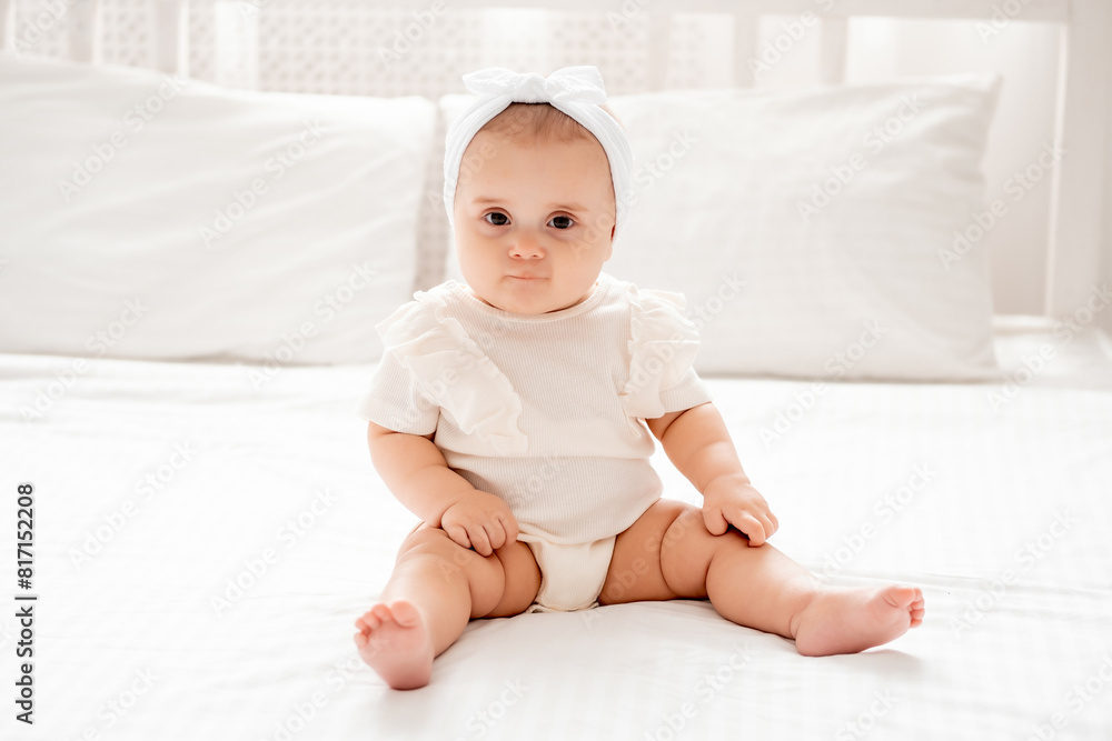 Cute newborn baby girl sitting on a bed in white clothes in a bright room, place for text, happy healthy baby close-up Cute newborn baby girl sitting on a bed in white clothes in a bright room, place for text, happy healthy baby close-up