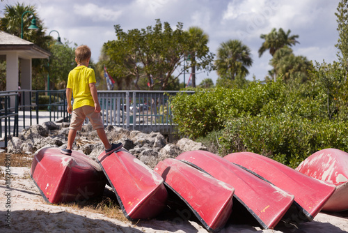 Kid boy standing on upturned boats on the river shore in the park on a sunny summer day. Child ready to sail. photo