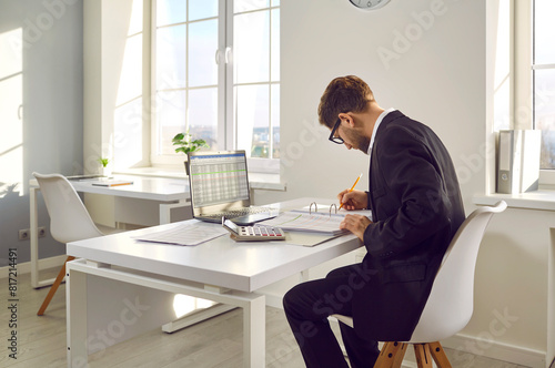 Accountant in a modern office workplace. Man in a suit sitting at his working desk, studying information in a thick paper binder and using business sheets on his laptop computer © Studio Romantic
