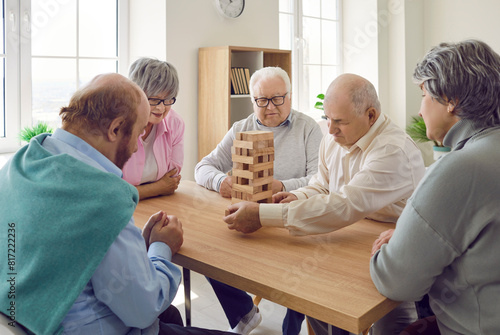 Group of senior men and women playing together with wooden building blocks for dementia therapy in nursing home enjoying activity sitting at the table. Leisure in retirement home concept.