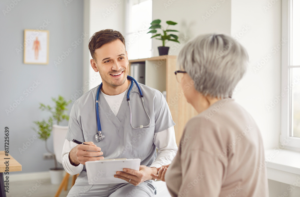 Friendly doctor gives informative medical consultation to elderly woman during clinic appointment. Smiling doctor with clipboard tells mature patient about treatment and explains examination results.