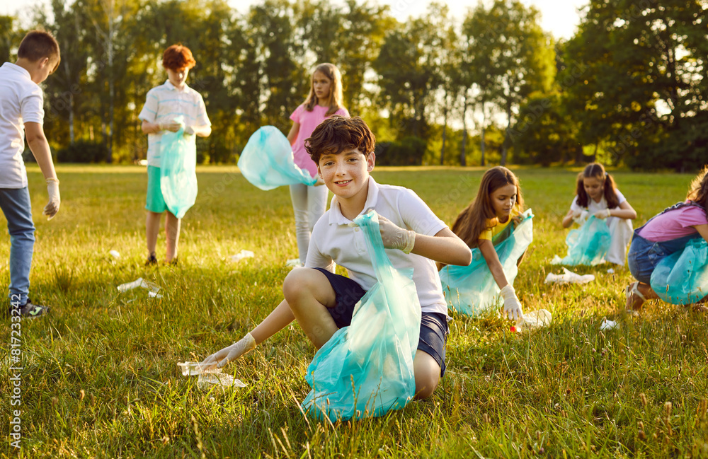 Fototapeta premium Save planet. Portrait of little boy who collects trash in garbage bag in forest or in park. Smiling preteen boy collects trash on green lawn in background of his peers who pick up used plastic.