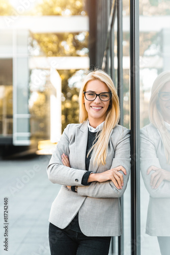 Intelligent confident blond businesswoman leaning on modern wall outside looking at camera.