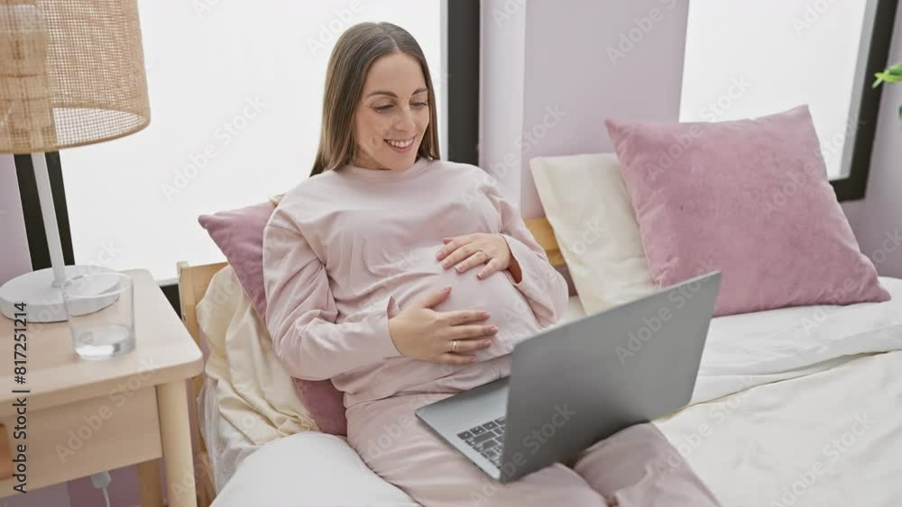 Smiling pregnant woman using laptop in bedroom, conveying expectancy and comfort at home.
