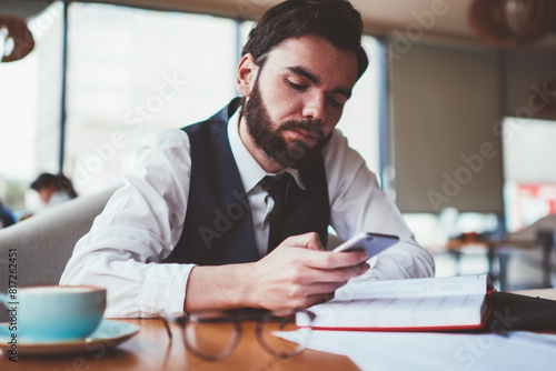 Bearded male blogger reading message from follover via telephone while creating new profile publication for social network  caucasian hipster guy updating notification on smartphone device indoors
