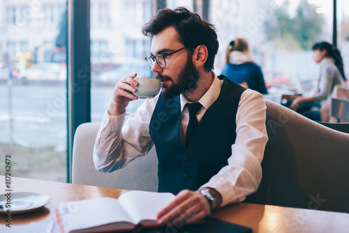 Fashionable hipster guy drinking hot coffee and looking at cafeteria window after reading interesting best seller, caucasian man pondering on literature during enjoying caffeine beverage indoors photo