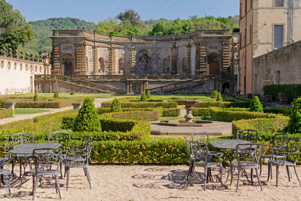 A beautiful summer park with figured trees and paths in the courtyard of an ancient villa, fenced with an ancient stone wall with sculptures, there is figured garden furniture made of metal