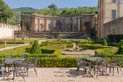 Obraz na płótnie A beautiful summer park with figured trees and paths in the courtyard of an ancient villa, fenced with an ancient stone wall with sculptures, there is figured garden furniture made of metal w salonie