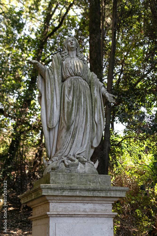 Life-size antique Roman sculpture of a woman in a tunic made of gray stone on a pedestal in a summer green park